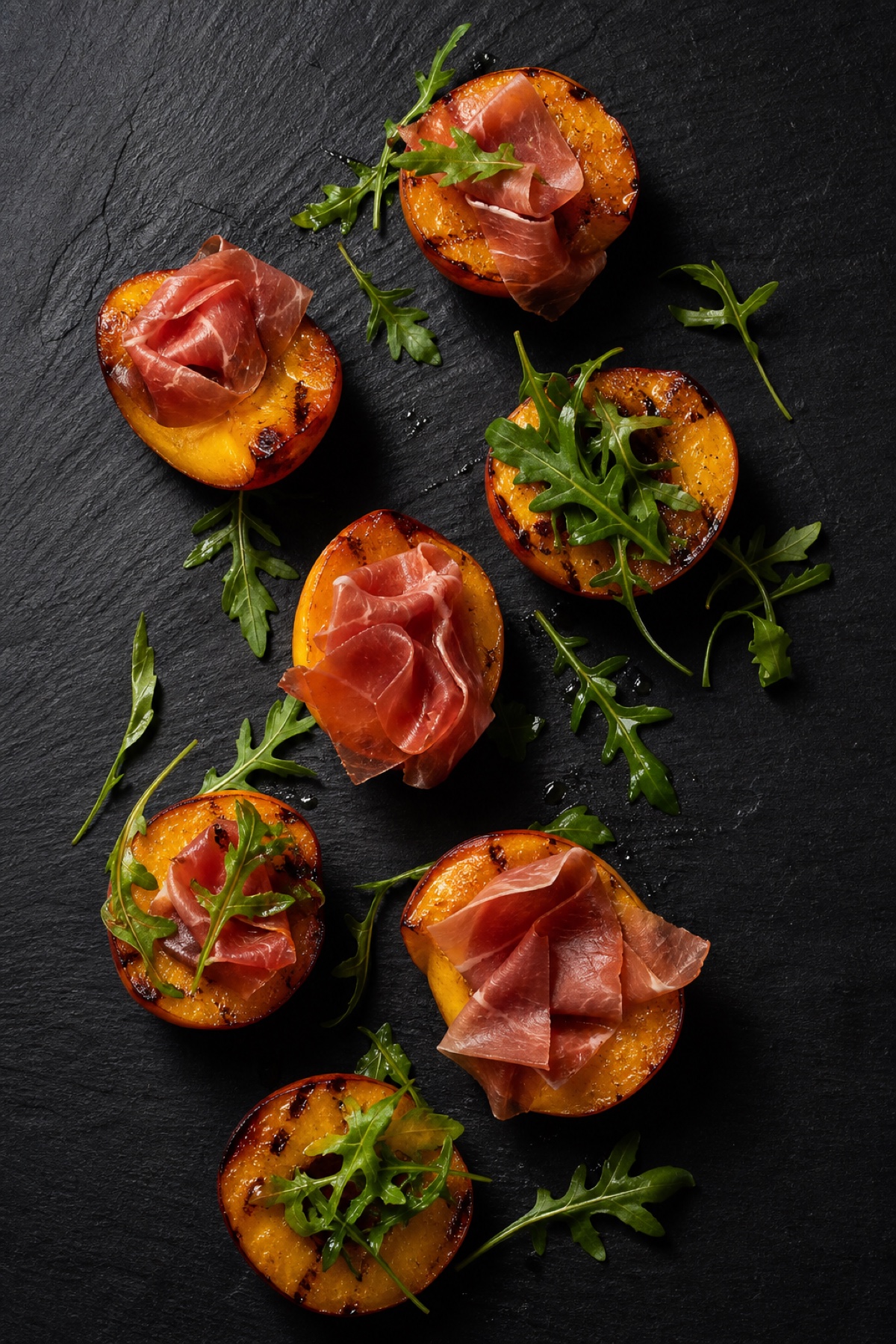 Grilled peach halves with draped prosciutto and rocket, overhead flat-lay on charcoal slate, gluten-free