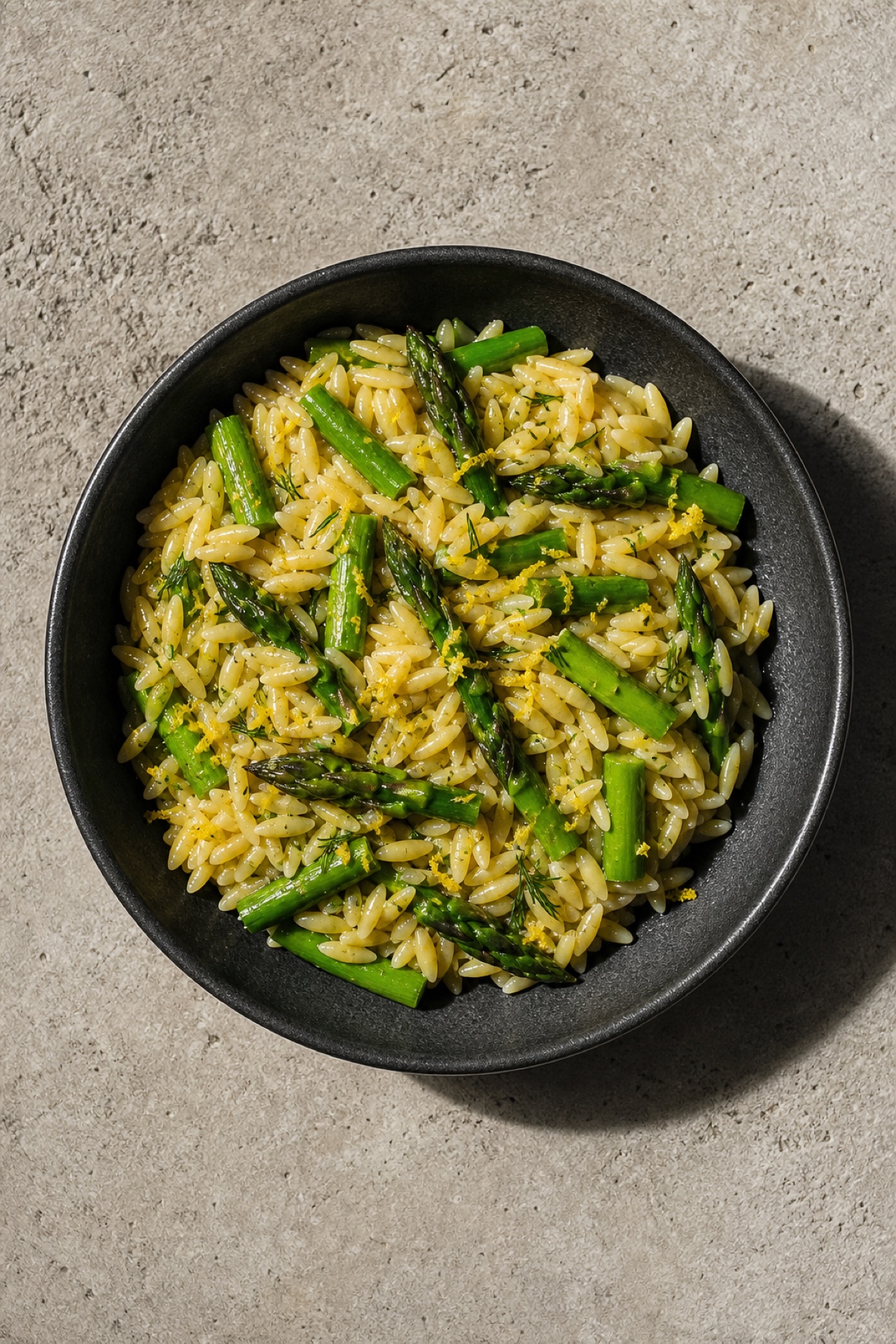 Lemon asparagus orzo in a dark bowl, overhead flat-lay on pale stone, vegan dairy-free
